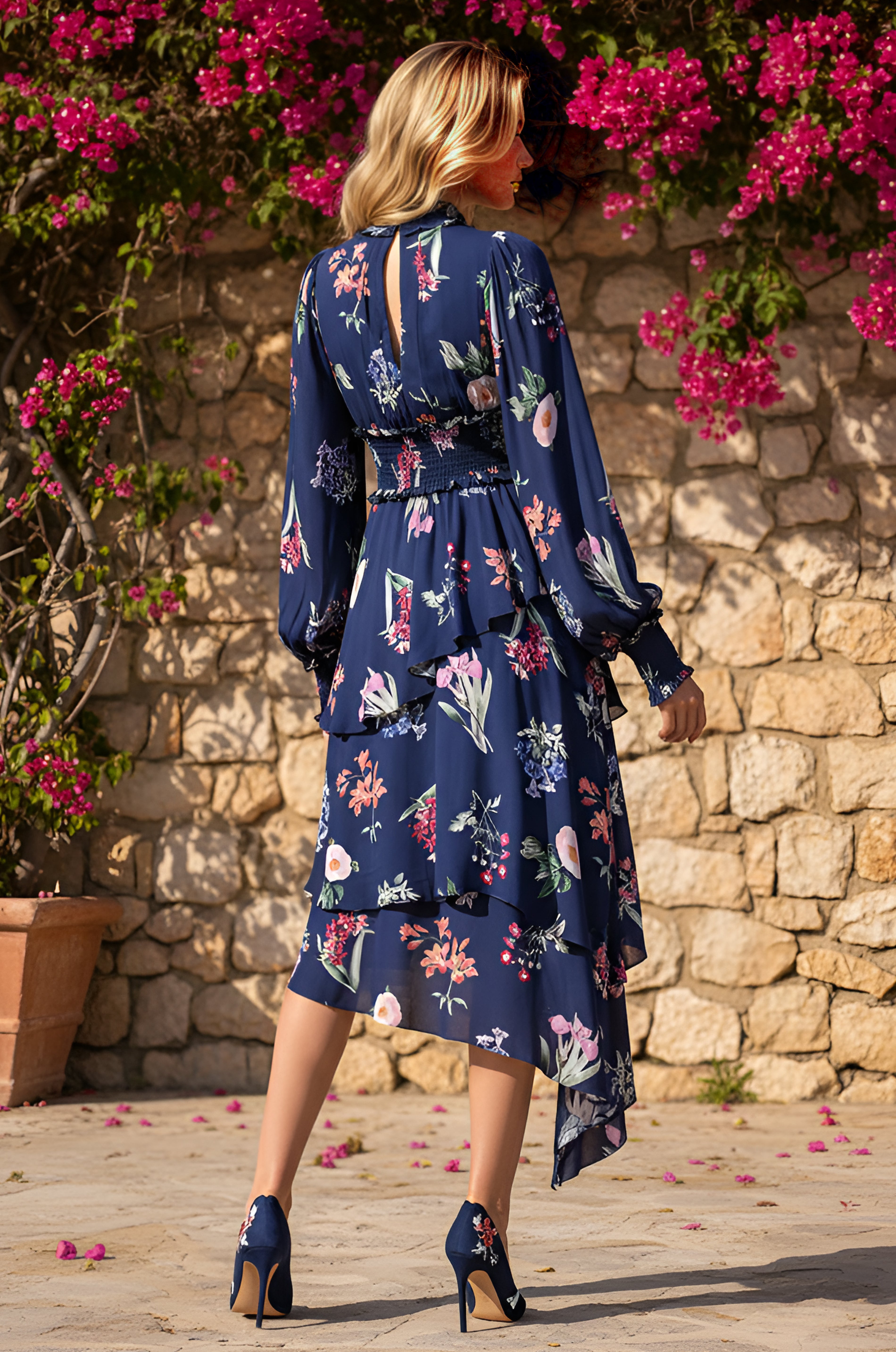 A woman in the Long-Sleeve Blue Floral Casual Midi Dress: Emery, featuring a smocked waistline and matching heels, stands outdoors on stone pavement, facing a stone wall topped with vibrant pink flowers.